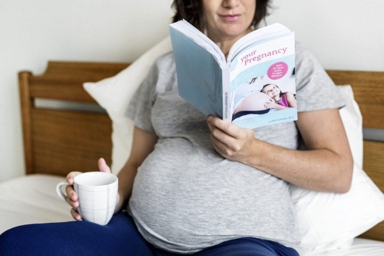 A pregnant person sits on a bed while reading a book titled "Your Pregnancy" and holding a white mug. The individual, clad in a grey t-shirt and blue pants, appears serene surrounded by pillows and a wooden headboard, embodying the essence of third trimester must-haves for comfort and relaxation.
