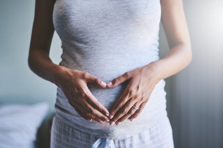 A person wearing a gray shirt and gray and white plaid pants is shown with hands placed over their stomach in a heart shape, showcasing first trimester must-haves. The setting appears to be indoors with soft light, with part of a bed visible in the background.