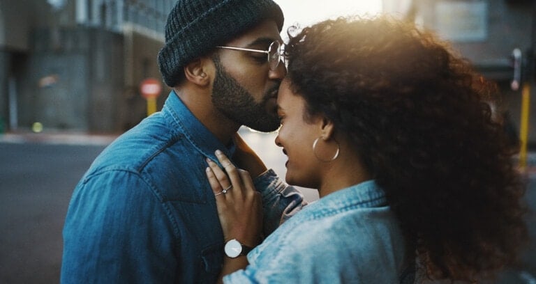 A man wearing glasses and a beanie kisses the forehead of a woman with curly hair. They stand close together, both dressed in denim jackets, in an outdoor urban setting with blurred buildings and street signs in the background, possibly sharing date night questions that deepen their connection.
