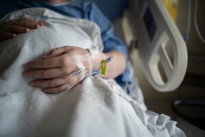 A close-up view of a person's hand resting on a blanket in a hospital bed. The hand has an IV inserted, secure with tape, highlighting the subtle difference between hep lock vs saline lock. The person is wearing a blue hospital gown, and the scene takes place within a hospital room.