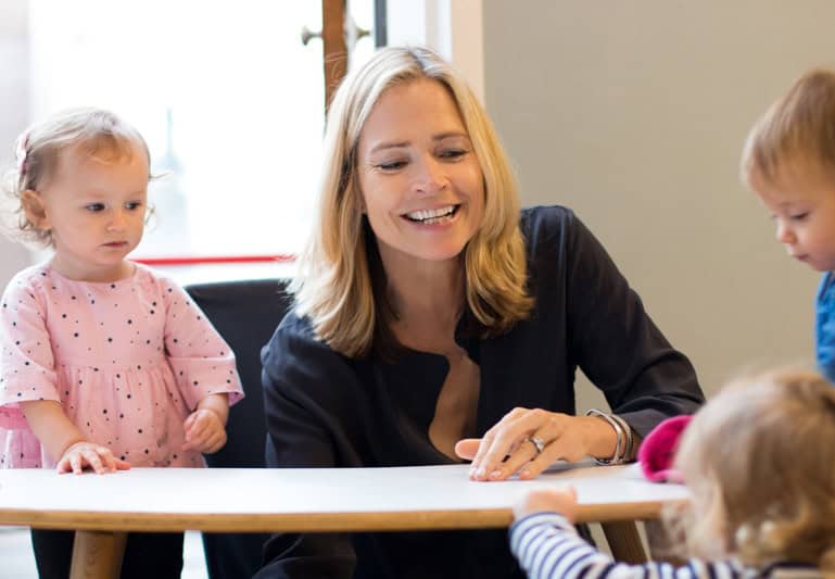 Janet Lansbury smiles while sitting at a round table with three young children, embodying the principles of respectful parenting. Two of the children stand by the table, while one child is partially visible in the foreground. The setting appears to be indoors with natural light.