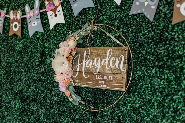 A decorative sign with the name "Hayden Elisabeth" displayed on a wooden plaque, adorned with flowers on a gold ring, hangs against a backdrop of green leaves. Perfect for a woodland baby shower, a banner featuring various animal faces is strung across the top of the image.