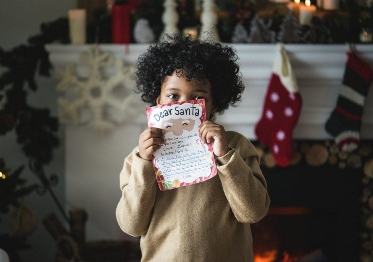 A child with curly hair stands in front of a fireplace, holding up a letter to Santa. The room is beautifully decorated for Christmas with stockings hanging on the mantel, candles, and festive ornaments—setting the perfect scene for dreaming about gifts for little boys.