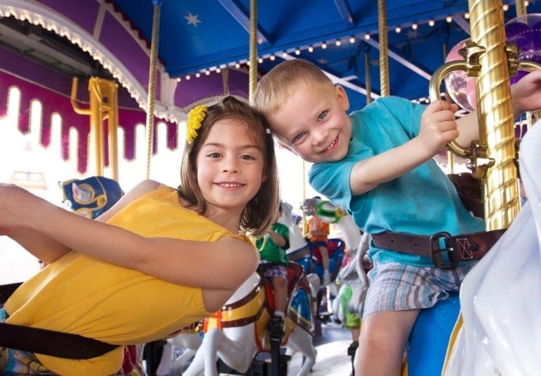 Two children are smiling while riding a carousel at Disney World. The girl on the left wears a yellow dress with a yellow flower in her hair, and the boy on the right wears a blue shirt and plaid shorts. Colorful carousel animals and lights are visible in the background, making it magical for any toddler.