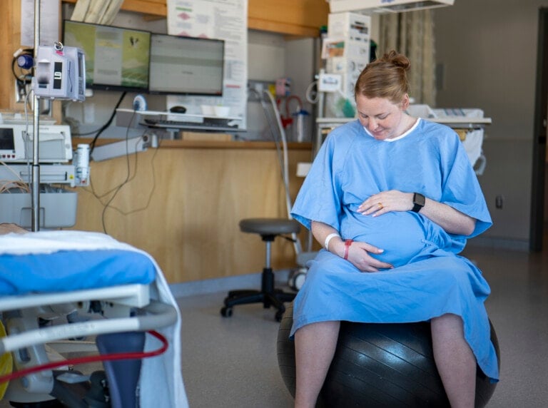 A pregnant woman in a blue hospital gown sits on an exercise ball in a medical room. She is looking down and placing her hands on her stomach. Hospital items, including medical equipment and a hospital bed, are visible in the background.