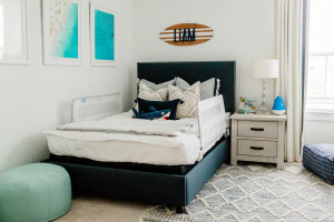 A bedroom with a neatly made bed featuring white and gray bedding, several pillows, and a protective bed rail. The walls have framed pictures and a decorative sign, perfect for a big boy room. A nightstand with a lamp and other items sits beside the bed, and a round green pouf is in the corner.