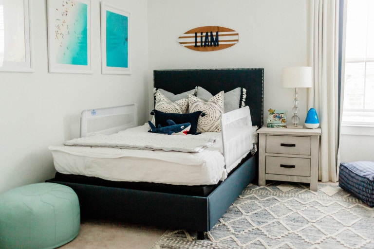 A bedroom with a neatly made bed featuring white and gray bedding, several pillows, and a protective bed rail. The walls have framed pictures and a decorative sign, perfect for a big boy room. A nightstand with a lamp and other items sits beside the bed, and a round green pouf is in the corner.