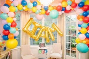 A balloon themed birthday party setup features a "Liam" sign in golden balloon letters surrounded by multi-colored balloons. Below, a high chair with a "1" decoration is placed between two white chairs in front of large windows with closed curtains.