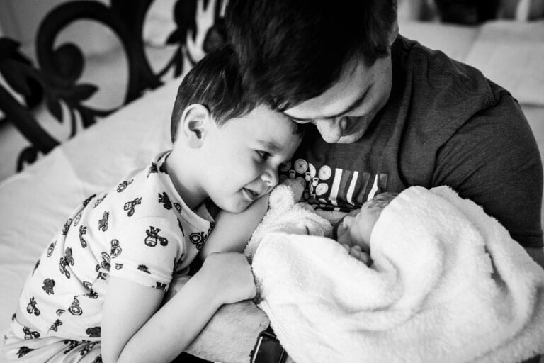 A black-and-white photo shows a man cradling a newborn wrapped in a blanket while a young boy, wearing pajamas, is leaning in close to look at the baby. They are all on a bed with ornate metal headboard details visible in the background—capturing the perfect moment for new baby gift ideas inspiration.