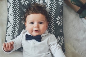 A baby with curly hair is lying down on a patterned cushion, wearing a white shirt and a navy blue bow tie. The cushion has a knitted snowflake design. The baby, who certainly deserves one of those unique boy names that start with A, is looking at the camera with a slight smile.