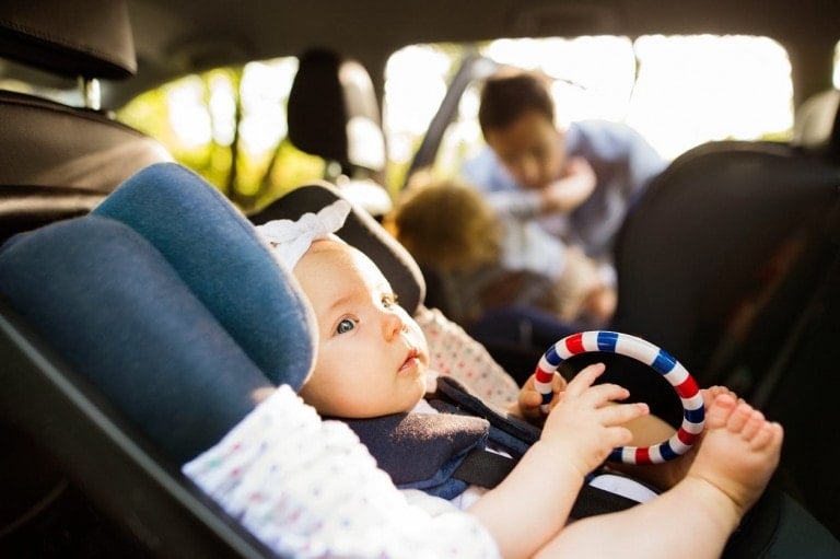 A baby is seated in a car seat inside a vehicle, holding a colorful ring toy, as sunlight filters through the windows, illuminating the baby's face. In the background, an adult interacts with another child—a serene moment of traveling with baby.