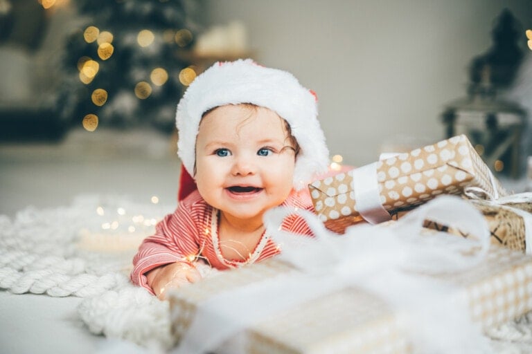 A baby wearing a Santa hat and red striped outfit is lying on a white rug with a few wrapped Christmas gifts in front. The background features holiday lights, a decorated Christmas tree, and festive birthday balloons. The baby is smiling and appears happy.