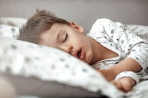 A young child with brown hair sleeps peacefully on a pillow, wearing white pajamas with small patterns. The child's mouth is slightly open, and he is lying on his side.