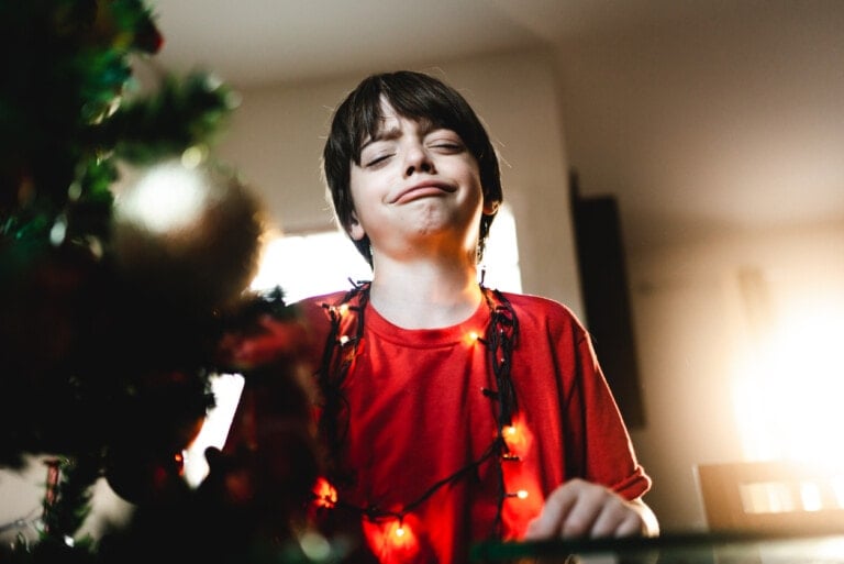 A young boy wearing a red shirt is tangled in Christmas lights, standing next to a decorated Christmas tree. He has a playful expression on his face, as if caught in the midst of a holiday meltdown. The background is softly lit by sunlight coming through a window.