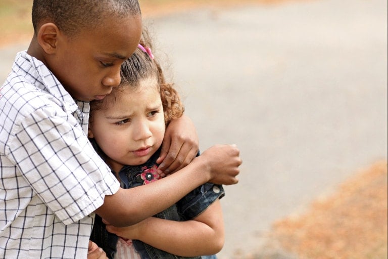A young boy and girl are standing outdoors. The boy, wearing a checkered shirt, is hugging the girl in a denim jacket. They both appear to be serious and thoughtful, perhaps in a moment of teaching empathy. The background is blurred with a paved path and fallen leaves.