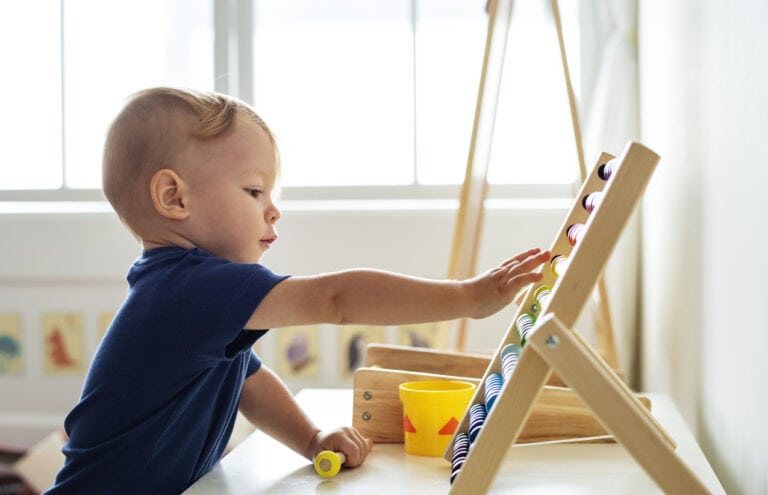 A young child, engrossed in infant activities, plays with a colorful abacus on a table in a bright room. The child, wearing a blue shirt, is focused on moving the beads. A yellow cup with a red triangle sits on the table next to the abacus, catering to their cognitive needs.