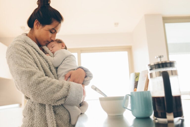 A woman wearing a bathrobe stands in a kitchen, gently holding and comforting a sleeping baby against her chest. On the counter nearby, there is a bowl with a spoon, a blue mug, and a French press coffee maker—essential items for many new mothers starting their day.