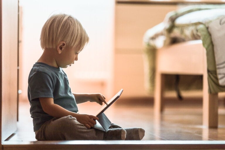 A young child with blonde hair is sitting on the floor, using a tablet device. Dressed in casual clothing, the child seems engrossed in one of the best reading apps for kids. The setting appears to be a softly lit bedroom with a bed in the background.