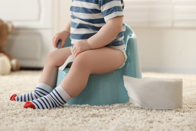 A toddler wearing a striped shirt and socks sits on a blue potty chair with a roll of toilet paper nearby on a carpeted floor, illustrating the challenges of potty training too early.