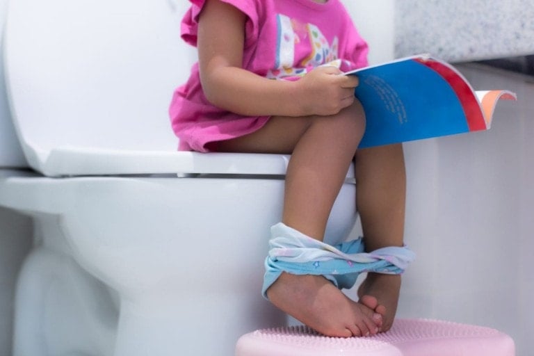 A child, in the midst of potty training, is seated on a white toilet with their underwear down, reading a book with a blue and red cover. Wearing a pink shirt and using a small pink step stool to rest their feet, the bathroom includes light-colored countertops that add to the calm atmosphere.