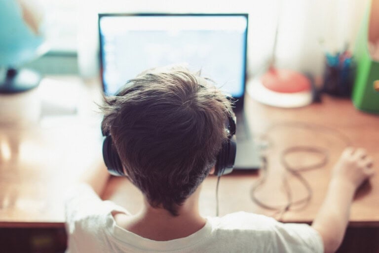 A child wearing over-ear headphones is sitting at a wooden desk, facing a computer screen. One hand is on a computer mouse, and the desk has various items including a lamp, globe, and stationery. The room is bright with natural light from a window.
