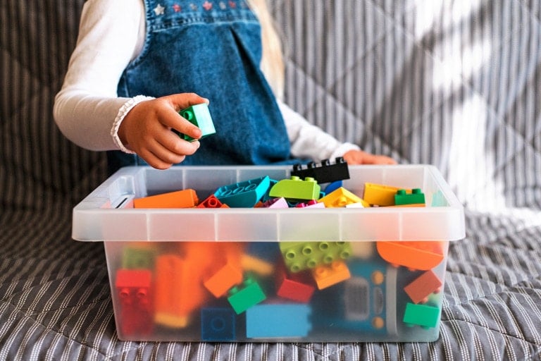 A child is sitting on a striped couch, holding a green building block over a transparent plastic container filled with various colorful building blocks. The child is wearing a white long-sleeve shirt and a blue denim dress, diligently engaged in toy storage.
