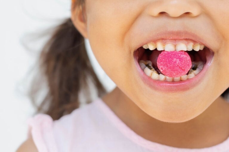 A close-up image shows a child with a bright pink candy resting on their tongue, smiling and displaying their teeth. The background is white and blurred, focusing attention on the child's mouth and candy, subtly hinting at the impact of sugar on kids.