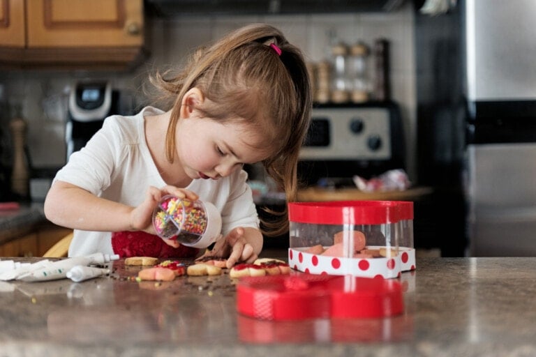A young child is decorating cookies at a kitchen counter as part of their Valentine's Day activities. The child is holding a container of colorful sprinkles and applying them to the cookies. The counter features a red and white polka dot box and decorating supplies, with the kitchen visible in the background.