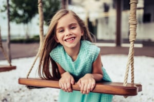 A young girl with long blonde hair is smiling, leaning forward on a wooden swing. She is wearing a light blue, textured dress. The swing has beige ropes, and there is a blurred background with greenery and a building. You could easily imagine her being called one of those cute nicknames for girls like "Sunshine.