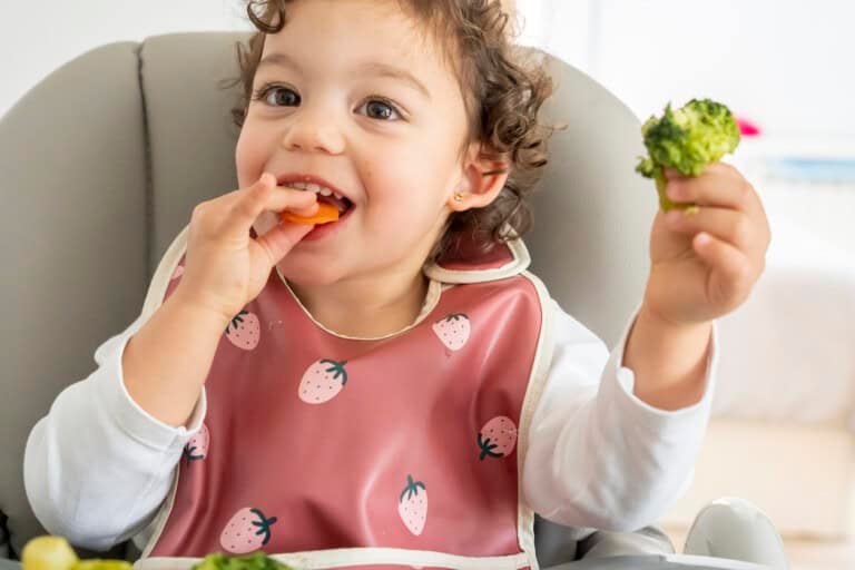 A young girl with curly hair sits in a high chair, wearing a bib with strawberry patterns. The child is holding a piece of broccoli in one hand and a carrot in the other, appearing to eat the carrot. A plate with more food is in front of them.