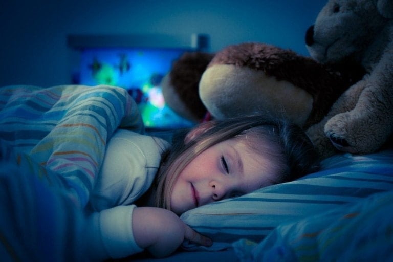A young child is sleeping in bed, covered with a striped blanket. The child is surrounded by stuffed animals, and a nightlight provides a soft blue glow in the background. The scene appears calm and peaceful, offering comfort against children night terrors.