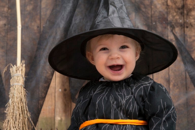 A baby dressed in a black witch costume with an orange belt smiles widely. The baby is wearing a large pointed witch hat. There is a straw broomstick on the left side of the image and a wooden background draped with dark fabric, adding to the enchantment of this scene perfect for any witchy names.