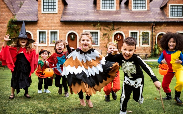 A group of children wearing Halloween costumes stand on a lawn in front of a house. The costumes include a witch, Little Red Riding Hood, a brown and white bird, a skeleton, and others. Some children hold orange buckets for trick-or-treating while discussing fun Halloween party games.