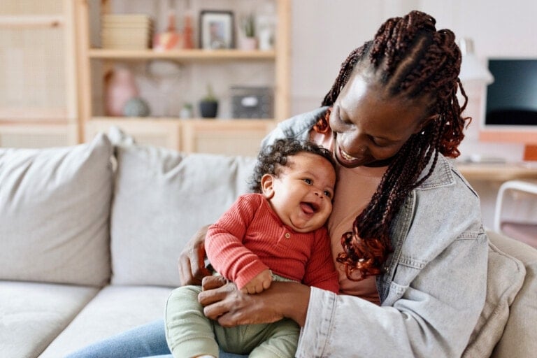 A woman with braided hair sits on a couch holding a smiling baby. The baby, adorably dressed in a red long-sleeve shirt and green pants, snuggles in her arms. Behind them is a bookshelf filled with various items and a desk, making the cozy living room feel complete. In case no one told you, those little moments matter most.