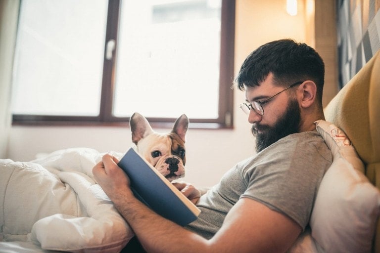 A man with a beard and glasses is leaning against a yellow headboard, reading baby books for your partner while sitting in bed. A French Bulldog, partially covered by a white duvet, is next to him, looking towards the camera. A window is visible in the background.