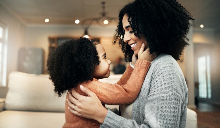 A woman and a child with curly hair sit on a couch indoors. The child, wearing an orange sweater, touches the woman’s face affectionately while she smiles and holds the child. This serene moment reflects mindful families, emphasizing the practice of mindfulness in parenting. The background shows a well-lit room with blurred details.