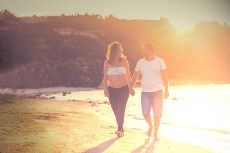 A couple holding hands walks along a beach during sunset, planning your babymoon as they savor the serene moment. The woman wears a white crop top and dark pants, while the man is dressed in a white T-shirt and shorts. The sun casts a warm glow over the scene, with the shoreline and hills visible in the background.