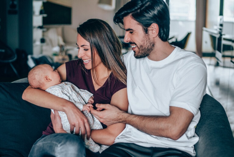 A woman and a man sitting on a couch are smiling and looking at a baby wrapped in a blanket. The woman is holding the baby, and the man is seated next to her, perhaps sharing advice for new parents. The background shows a blurry interior of a home.