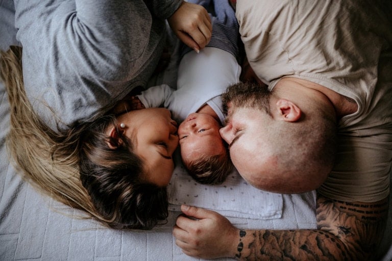 A newborn baby lying on a blanket is flanked by two adults, one on either side. The person on the left is kissing the baby's forehead while the person on the right looks at the baby. They are all lying on their sides on a bed, perhaps discussing their postpartum plan for this new chapter in life.