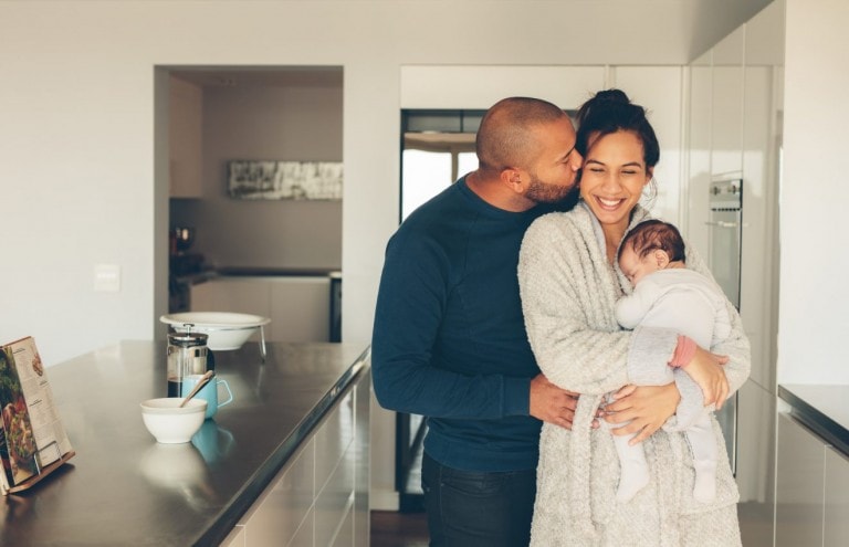 In a well-lit, modern kitchen, a man kisses a woman’s temple as she smiles and holds a baby. Nearby, on the counter, sit a coffee maker, cookbook, bowl, and mug. This tender moment reflects the couple's postpartum love languages amid their new journey of parenthood.