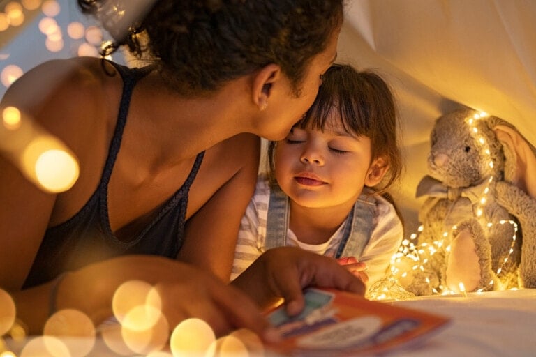 A woman kisses a young child on the forehead while lying in a tent decorated with fairy lights. The child, holding a classic book, appears relaxed with eyes closed. A stuffed rabbit wrapped in lights is next to the child.
