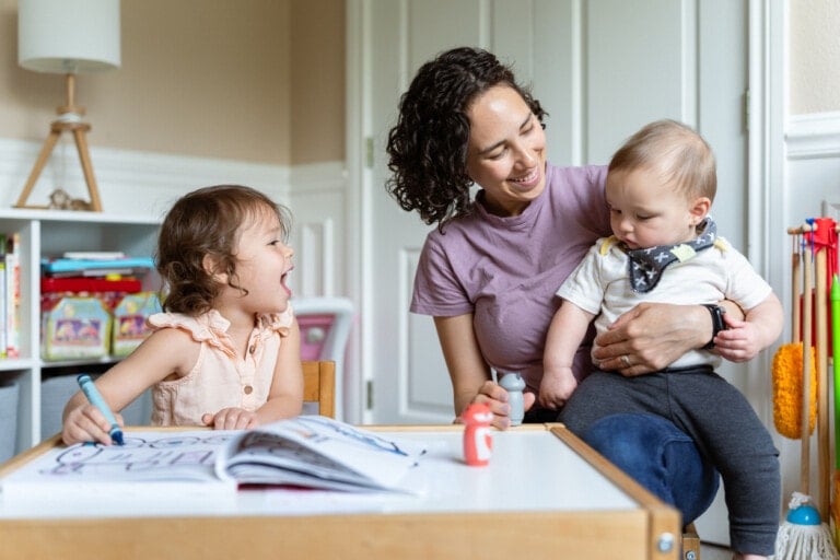 A woman sits with a happy toddler on her lap, smiling at a young girl who is drawing with markers at a small table. The room, filled with children's furniture, toys, and bookshelves in the background, perfectly captures the joyful essence of toddler play.