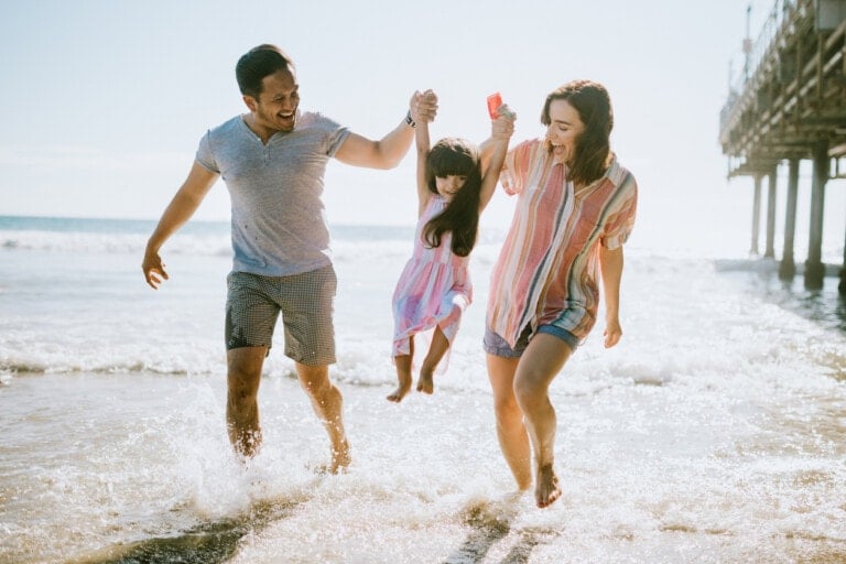A man and a woman are walking on the beach, holding the hands of a young girl between them. The girl is being lifted off the ground and is smiling. Waves gently crash around their feet, and a pier is seen in the background—a perfect family vacation idea.