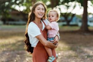 A woman with long brown hair and a white shirt under a rust-colored overall holds her 17-month-old, who is in a pink shirt, as they enjoy the outdoors. The toddler looks towards the camera with curiosity. Blurred trees create a serene backdrop, capturing this beautiful moment of motherhood. A beautiful and happy woman of Pacific Islander descent holds her curious one year old Eurasian son on her hip and affectionately talks with the child as they spend quality time together outside at a park in Hawaii.
