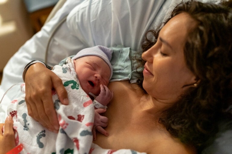 A woman lying in a hospital bed smiles while holding a newborn baby wrapped in a blanket with a foot print pattern. The baby, wearing a tiny hat, rests against the woman's chest as she experiences the joy Henci Goer often describes in her writings on childbirth.