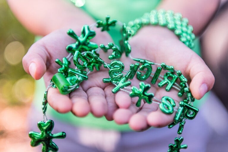 Close-up of a person holding several green metallic necklaces in their hands. The necklaces include shamrock shapes and letters, making it perfect for a St. Patrick's Day activity. The background is blurred, focusing on the person’s hands and the festive items they are holding.