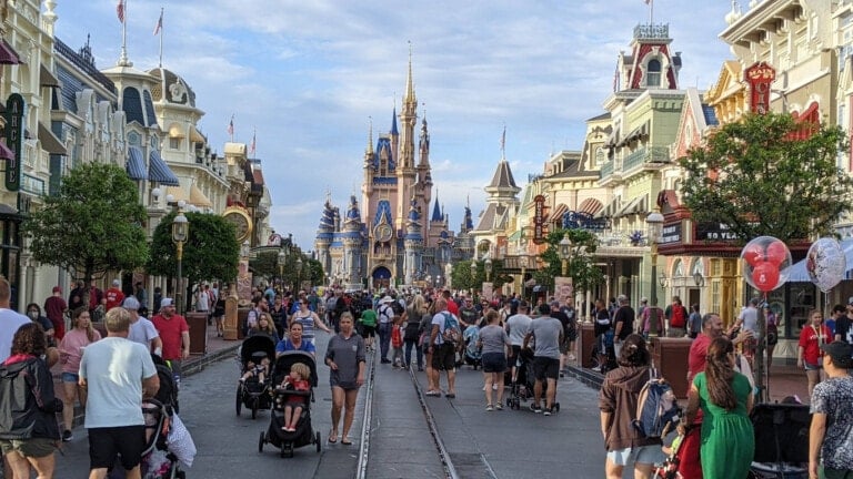 A bustling street at Disney World filled with visitors, some pushing strollers or carrying balloons. At the end of the street, a large, ornate castle with spires can be seen. Buildings with colorful facades line both sides of the path, creating a magical experience for families exploring Disney World with babies in tow.
