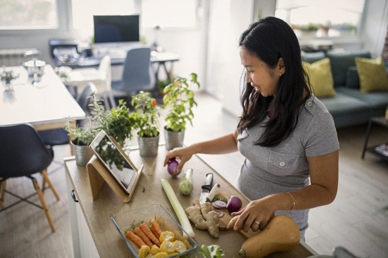 A woman standing in a kitchen using a tablet for first trimester recipes. She is chopping vegetables, including carrots and eggplants, on a cutting board. There are herbs and other ingredients on the counter, and a spacious living area is visible in the background.