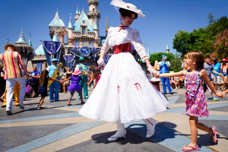 A woman dressed as a character in a white dress, wide-brimmed hat, and red belt holds hands and dances with a young girl in a colorful dress at an outdoor theme park during their Disney vacation with kids, with onlookers and characters in the background. A castle structure is visible behind them.