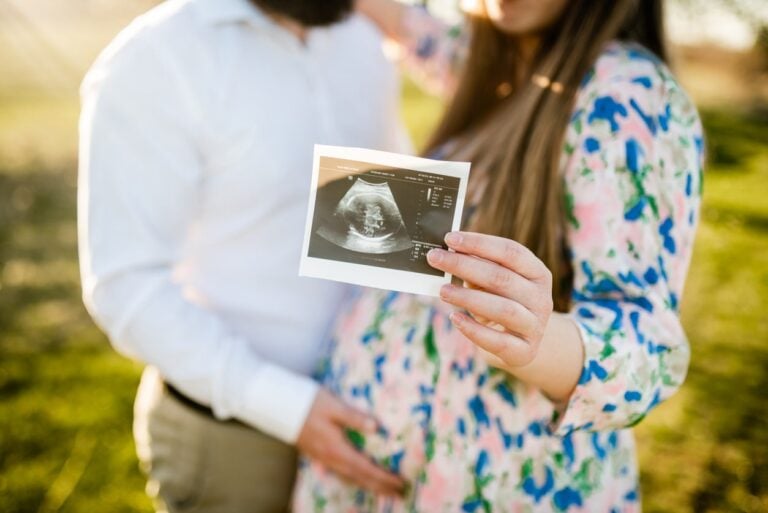 A couple stands outdoors, with the woman's hand holding a sonogram image in the foreground. The man, wearing a white shirt, gently holds the woman's pregnant belly. The woman is in a floral dress, embodying love and hope that echo timeless pregnancy quotes. The background is grassy and sunlit.
